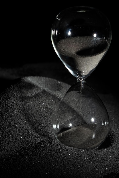 Foto orologio di sabbia in piedi su sabbia nera con ombra di silhouette su sfondo nero orologi di sabbia neri mostrano più tempo scadenza prolungata gestione del tempo speranza concetto ora orologio di vita di vetro che passa