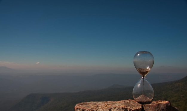 A hourglass (with falling sand) on a cliff rock with forest mountain in background