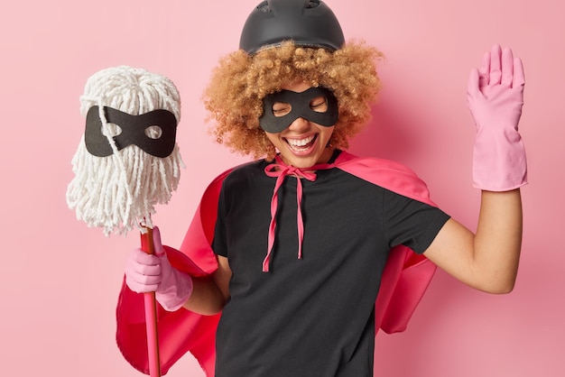 House cleaning again positive curly haired woman dressed like\
superhero wears helmet eyemask rubber gloves holds mop going to\
wash floor in room has happy expression isolated over pink\
background