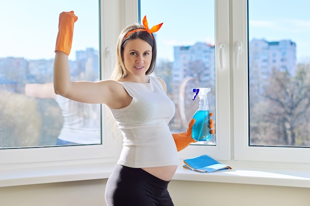 House cleaning, young beautiful pregnant woman in gloves with rag and detergent posing, looking at camera, near clean washed window in room