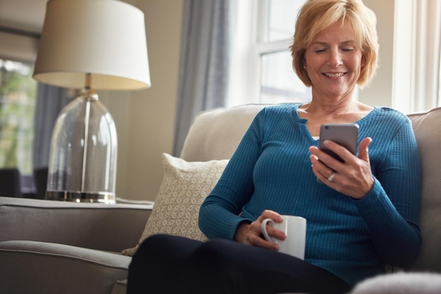 House coffee and old woman on sofa with smartphone typing or connection with social media in lounge Happy senior person apartment or mature lady with cellphone internet or relax with digital app