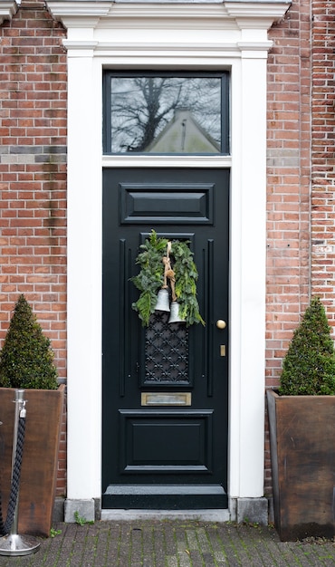 House facade with vintage front door decorated with christmas wreath with bells. Black vintage door with festive christmas decor. Porch European style in brick house.