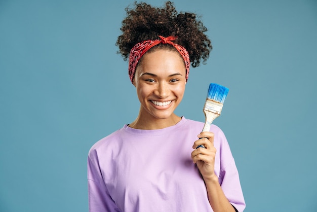 House improvement and renovation concept. Happy curly haired woman holding dirty paint brush and preparing to the repair while moving in new flat. Female painter