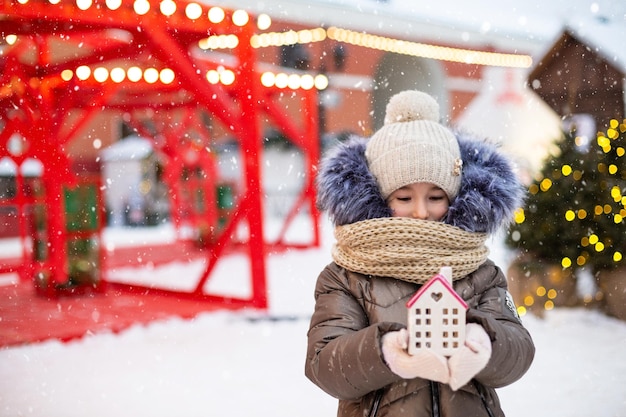 House miniature cottage in hands of girl wearing mittens and\
warm clothes outdoor in snow family values purchase housing\
relocation mortgage cozy home christmas new year booking