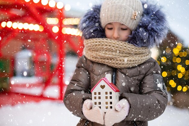 House miniature cottage in hands of girl wearing mittens and\
warm clothes outdoor in snow family values purchase housing\
relocation mortgage cozy home christmas new year booking