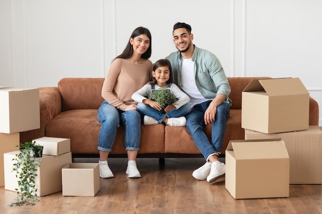 House Move And Renovation Concept. Portrait of happy loving family of three people sitting on the couch in new empty apartment, posing and looking at camera, parents embracing girl holding plant