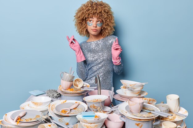 Household and home hygiene routine concept thoughtful woman\
wears festive silver dress rubber protective gloves surrounded by\
pile of dirty dishes with food leftovers poses indoor on blue\
background