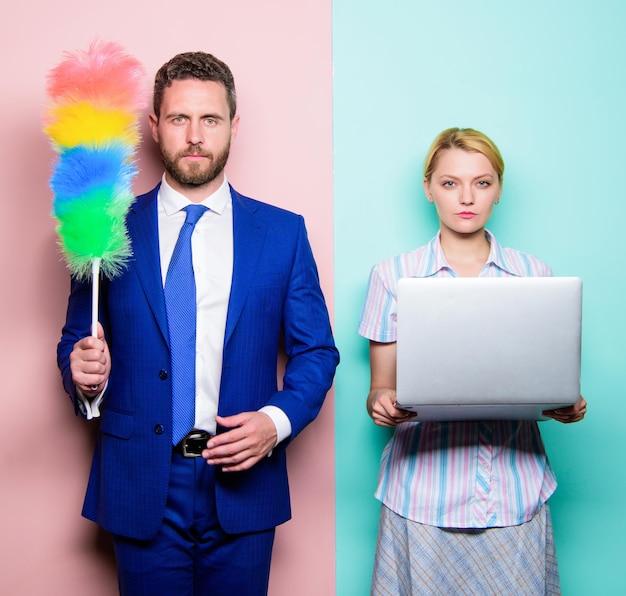 Househusband and female boss. Businessman and housewife. Family couple. Husband stand with dust brush while wife holding laptop. Couple of handsome man and pretty woman. Housekeeping or business.