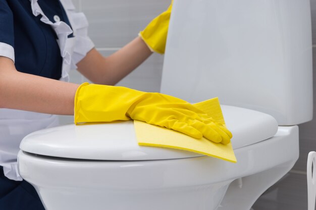 Housekeeper cleaning a white toilet lid wiping it down with a yellow cloth with her gloved hands, close up view