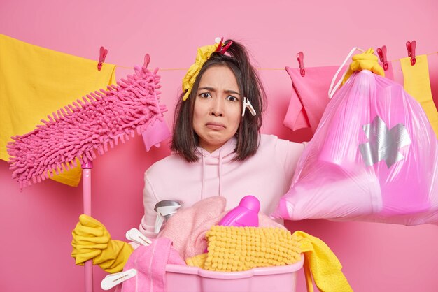 Housekeeping and cleaning service concept displeased tired\
asian housekeeper holds mop and garbage bag wears sweatshirt and\
rubber gloves collects laundry in basket busy washing\
everything