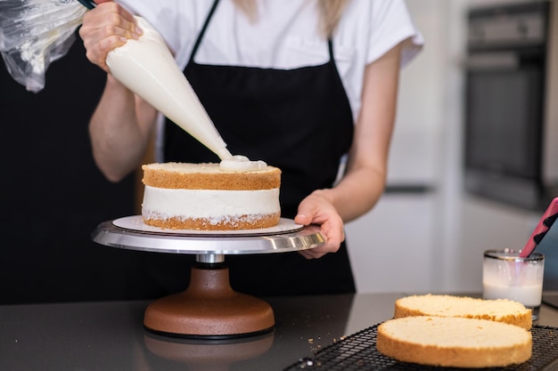 Домохозяйка в черном фартуке с помощью конуса для выпечки смазывает белым кремом свежие слои торта