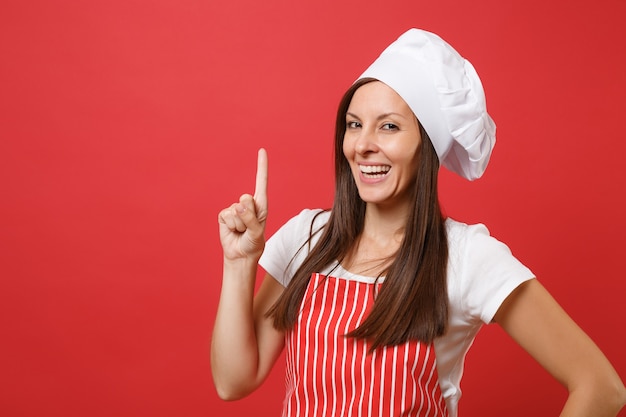 Housewife female chef cook or baker in red striped apron, white t-shirt, toque chefs hat isolated on red wall background. Close up portrait of housekeeper brunette woman. Mock up copy space concept.