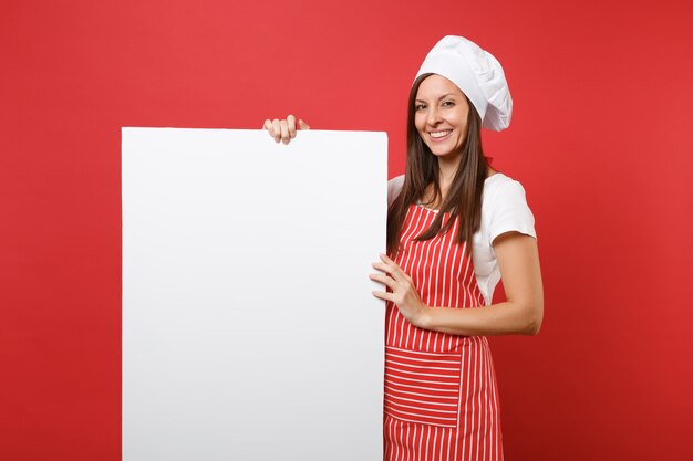 주부 여성 셰프는 줄무늬 앞치마, 흰색 티셔츠, 빨간색 배경에 격리된 토크 셰프 모자를 쓰고 제빵사를 요리합니다. 여자는 판촉 콘텐츠를 위해 큰 흰색 빈 광고판을 들고 있습니다. 복사 공간 개념을 비웃습니다.