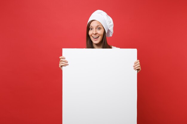縞模様のエプロン、白いTシャツ、赤い背景で隔離のトーク帽のシェフの帽子の主婦女性シェフ料理パン屋。女性はプロモーションコンテンツのために大きな白い空白の看板を持っています。コピースペースの概念をモックアップします。