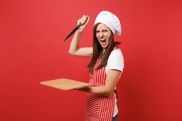 Housewife female chef cook or baker in striped apron white t-shirt, toque chefs hat isolated on red wall background. Angry screaming woman hold wooden cutting board, knife. Mock up copy space concept.