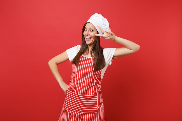 Housewife female chef cook or baker in striped apron white t-shirt toque chefs hat isolated on red wall background. Beautiful housekeeper woman showing victory sign at eye. Mock up copy space concept.