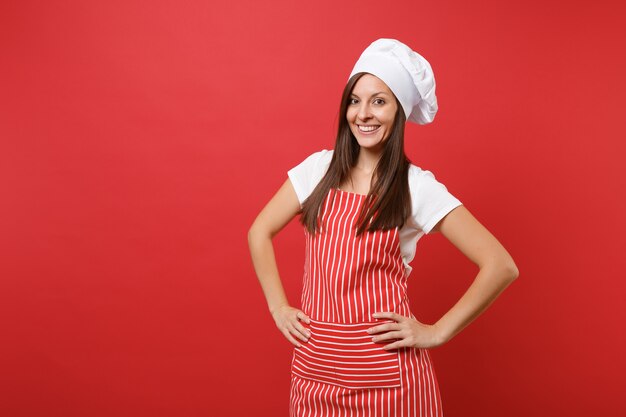 Housewife female chef cook or baker in striped apron, white t-shirt, toque chefs hat isolated on red wall background. Beautiful housekeeper woman standing with arms akimbo. Mock up copy space concept.