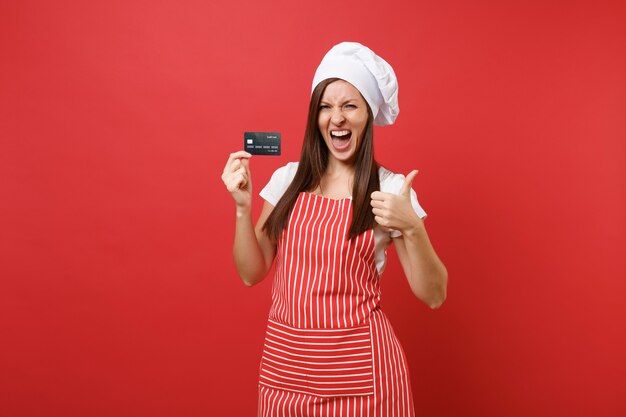 Housewife female chef cook baker in striped apron white t-shirt toque chefs hat isolated on red wall background. Excited woman hold in hand credit bank card, cashless money. Mock up copy space concept