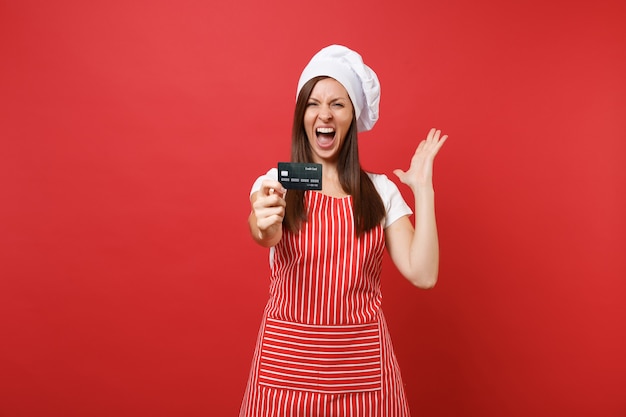 Housewife female chef cook baker in striped apron white t-shirt toque chefs hat isolated on red wall background. Excited woman hold in hand credit bank card, cashless money. Mock up copy space concept