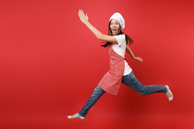 Housewife female chef cook or baker in striped apron white t-shirt, toque chefs hat isolated on red wall background. Full length portrait housekeeper woman jumping high up. Mock up copy space concept.