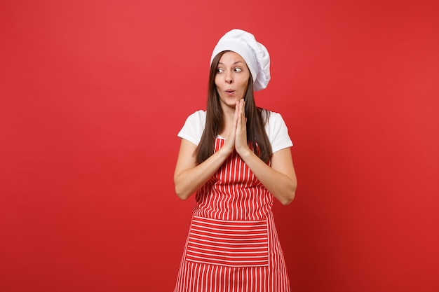 縞模様のエプロンの白いTシャツ、赤い壁の背景に分離されたトーク帽のシェフの帽子の主婦の女性シェフの料理人またはパン屋。楽しいかわいい家政婦の女性が期待して腕を組んだ。コピースペースの概念をモックアップします。