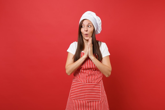 Foto cuoco o fornaio femminile della casalinga in maglietta bianca del grembiule a strisce, cappello del cuoco unico di toque isolato sul fondo rosso della parete. la donna carina e divertente della governante ha piegato le braccia in attesa. deridere il concetto dello spazio della copia.