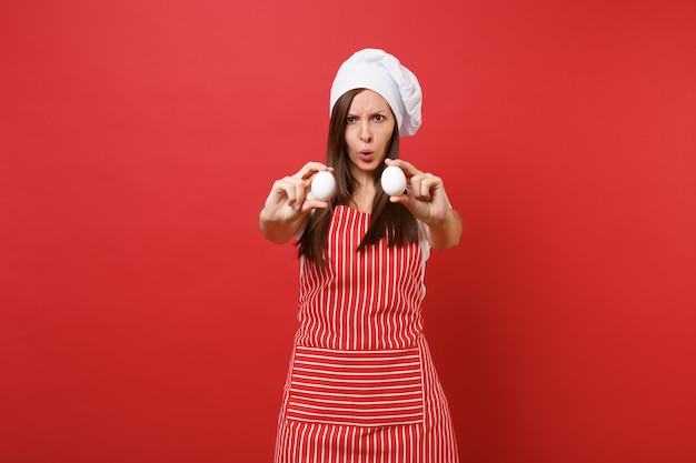 주부 여성 셰프는 줄무늬 앞치마, 흰색 티셔츠, 빨간 벽 배경에 격리된 토크 셰프 모자를 쓰고 제빵사를 요리합니다. 재미있는 가정부 여자는 두 개의 닭고기 달걀을 손에 쥐고 있습니다. 복사 공간 개념을 비웃습니다.