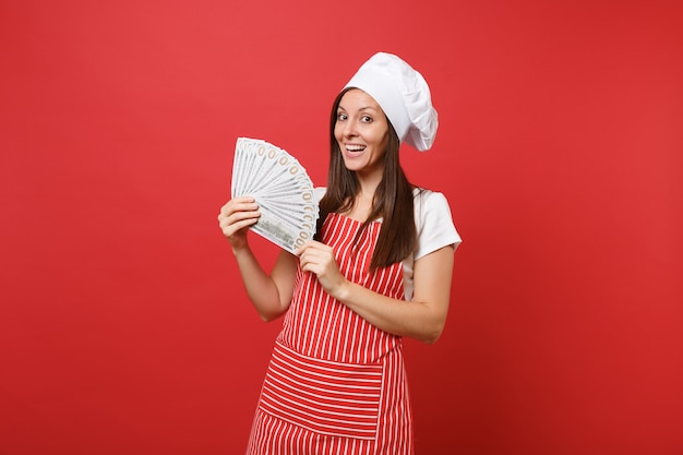 縞模様のエプロン、白いTシャツ、赤い壁の背景に分離されたトーク帽のシェフの帽子の主婦女性シェフ料理パン屋。たくさんのドル紙幣の現金を持っている幸せな女性。コピースペースの概念をモックアップします。