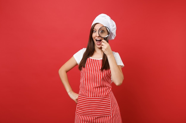 Housewife female chef cook or baker in striped apron white t-shirt toque chefs hat isolated on red wall background. Housekeeper woman hold and look through magnifying glass. Mock up copy space concept