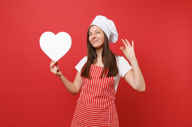 Foto cuoco o fornaio femminile della casalinga in grembiule a strisce, t-shirt bianca, cappello da cuoco unico del toque isolato sul fondo rosso della parete. la donna sorridente della governante tiene l'area di lavoro vuota del cuore. deridere il concetto dello spazio della copia.