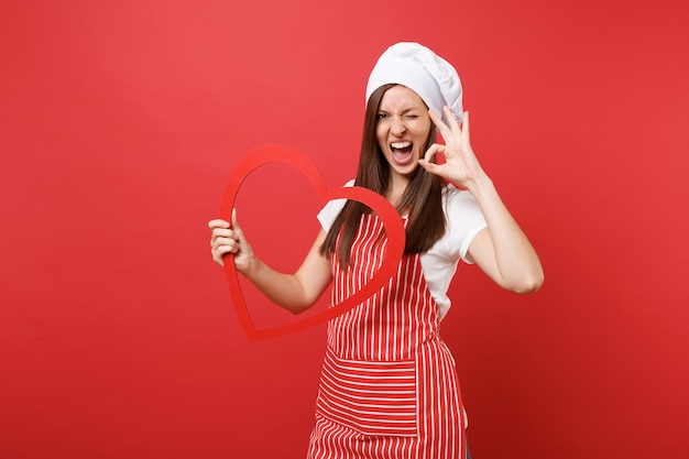 縞模様のエプロン、白いTシャツ、赤い壁の背景に分離されたトーク帽のシェフの帽子の主婦女性シェフ料理人またはパン屋。木製の赤いハートを持って笑顔の家政婦の女性。コピースペースの概念をモックアップします。
