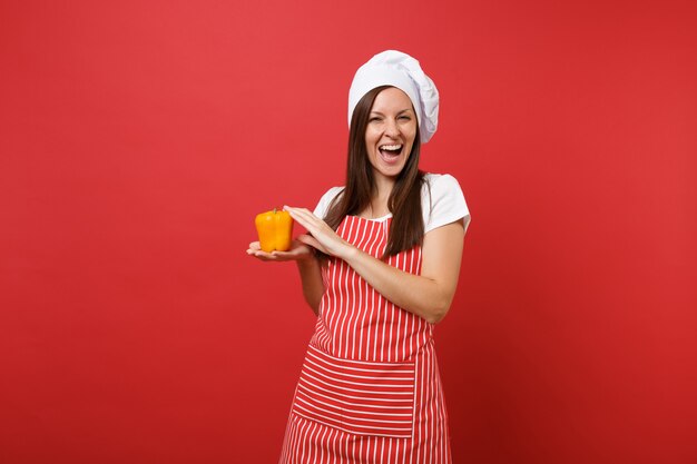 Housewife female chef cook or baker in striped apron, white t-shirt, toque chefs hat isolated on red wall background in studio. Smiling woman hold in hand yellow pepper. Mock up copy space concept.