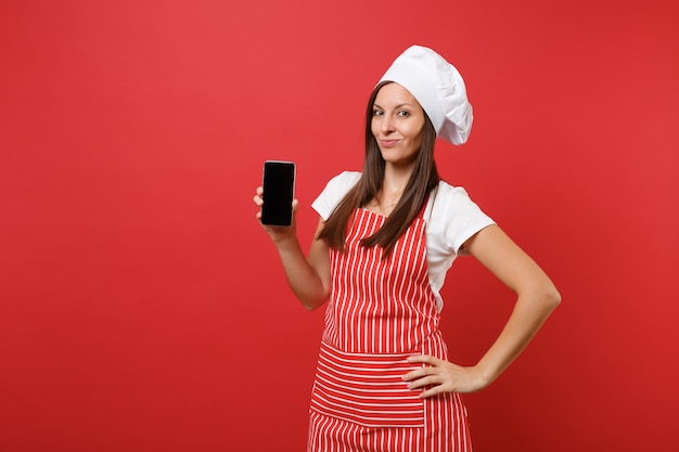 Housewife female chef cook or baker in striped apron white t-shirt toque chefs hat isolated on red wall background. Woman hold cellphone blank screen for promotional content Mock up copy space concept