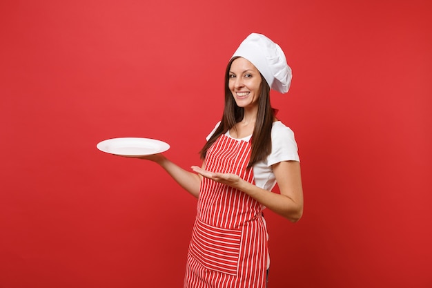 Housewife female chef cook or baker in striped apron, white t-shirt, toque chefs hat isolated on red wall background. woman hold empty blank round plate with place for food. mock up copy space concept