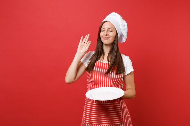 Housewife female chef cook or baker in striped apron, white t-shirt, toque chefs hat isolated on red wall background. Woman hold empty blank round plate with place for food. Mock up copy space concept