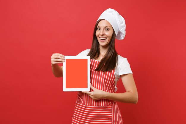 붉은 벽 배경에 격리된 줄무늬 앞치마 흰색 티셔츠 토크 셰프 모자를 쓴 주부 여성 요리사 또는 제빵사. 여성은 판촉 콘텐츠를 위해 태블릿 PC 빈 화면을 잡고 복사 공간 개념을 조롱합니다.