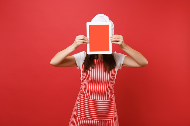 Housewife female chef cook or baker in striped apron white t-shirt toque chefs hat isolated on red wall background. Woman hold tablet pc blank screen for promotional content Mock up copy space concept