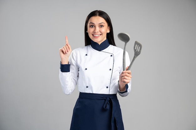 Housewife female chef cook or baker in uniform holding finger up and having idea isolated on grey wall background. Stock photo