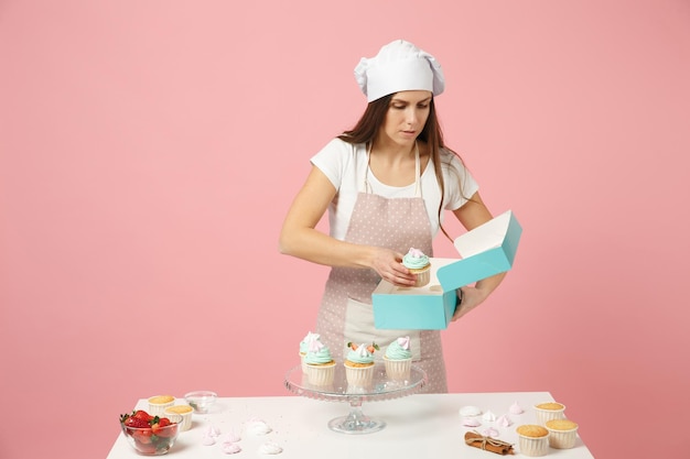 Housewife female chef cook confectioner or baker in apron white t-shirt, toque chefs hat packaging cake cupcake at table isolated on pink pastel background in studio. Mock up copy space food concept.