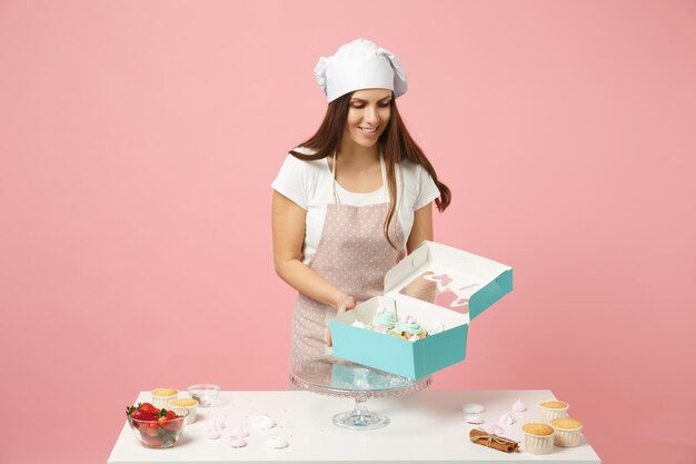 Housewife female chef cook confectioner or baker in apron white\
t-shirt, toque chefs hat packaging cake cupcake at table isolated\
on pink pastel background in studio. mock up copy space food\
concept.