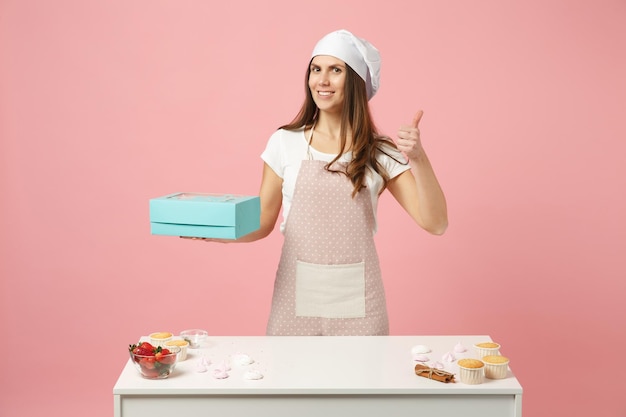 Housewife female chef cook confectioner or baker in apron white\
t-shirt, toque chefs hat packaging cake cupcake at table isolated\
on pink pastel background in studio. mock up copy space food\
concept.