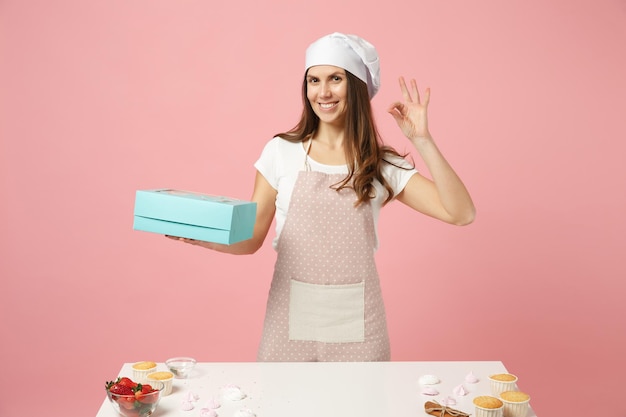 Housewife female chef cook confectioner or baker in apron white\
t-shirt, toque chefs hat packaging cake cupcake at table isolated\
on pink pastel background in studio. mock up copy space food\
concept.