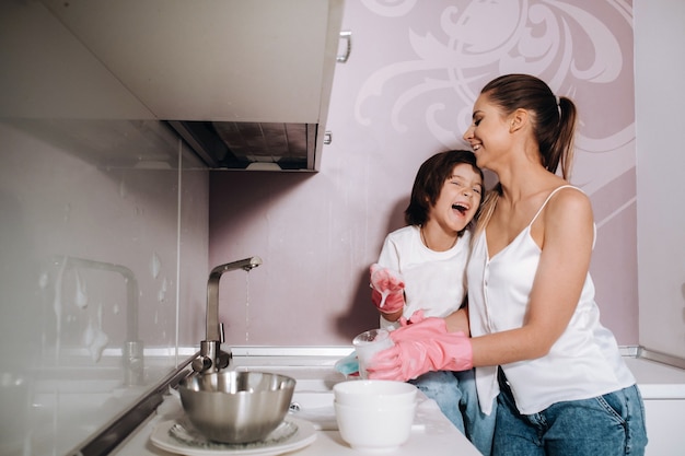Housewife mom in pink gloves washes dishes with her son by hand in the sink with detergent. A girl in white and a child with a cast cleans the house and washes dishes in homemade pink gloves.