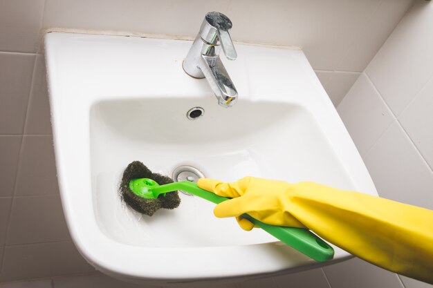 Housewife wearing yellow rubber glove and using brush cleaning dirty basin in the toilet