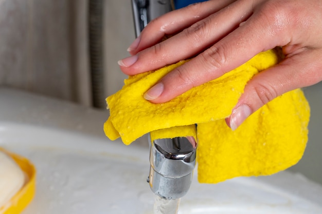주부는 욕실에서 수도꼭지를 청소하기 위해 젖은 천을 노란색으로 만듭니다. 집 청소 및 청결 유지의 개념
