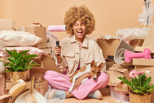 Housing repair and moving in concept positive curly woman holds
paintr roller going to redecorate walls in room of new apartment
poses with dog collects personal belongings in carton
containers