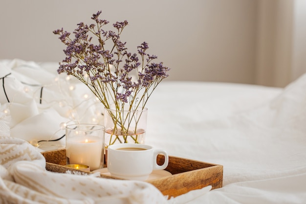 Houten dienblad met koffie en kaarsen met bloemen op bed. Witte beddengoed lakens met gestreepte deken en kussen. Ontbijt op bed. Hygge-concept.