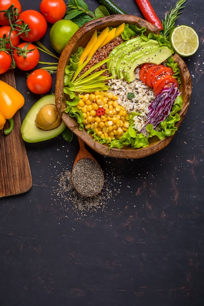 Foto houten kom met kikkererwten, avocado, wilde rijst, quinoa, paprika, tomaten, greens, kool, sla op donkere stenen tafel en houten lepel met chiazaden. bovenaanzicht met copyspace.