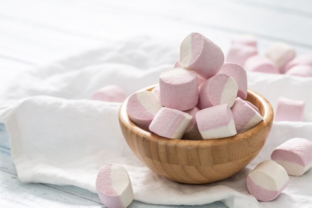 Houten kom vol roze en witte marshmallows met wat verspreid op een wit tafelkleed.