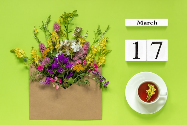 Houten kubussen kalender 17 maart. Kopje kruiden thee, kraft envelop met multi gekleurde bloemen op groene achtergrond